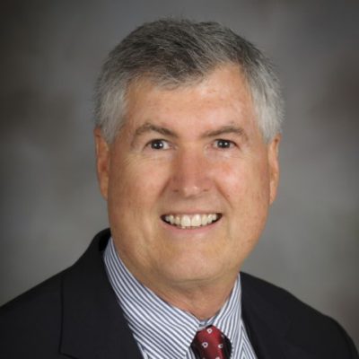 Dr. Price smiles for the camera, relaxed and happy, wearing a blue plaid shirt which is open over a t shirt. He is poised and confident